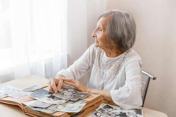 Ältere Frau, die zu Hause ein schwarzweißes altes Fotoalbum betrachtet. Die Person, die ihr eigenes Foto betrachtet, stammt aus den Jahren 1963, 1965. Sie lächelt, wenn sie sich daran erinnert, wie jung sie war, und schaut aus dem Fenster. - Foto, Bild