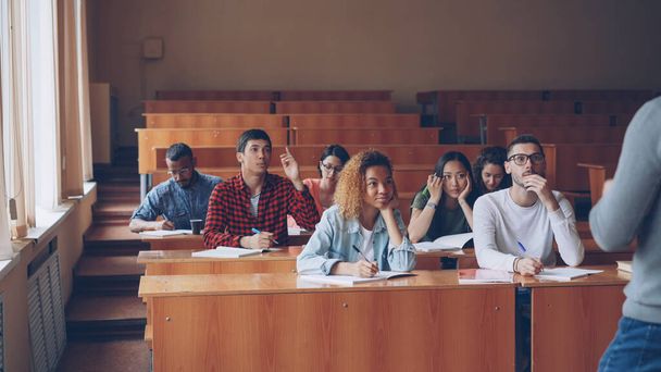 Multi-ethnic group of students is answering teachers questions, smart young people are talking and smiling sitting in tables while teacher is asking them checking knowledge. - 写真・画像
