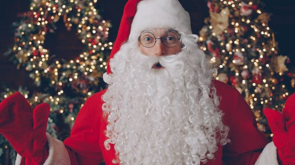 Portrait of man in traditional Santa Claus costume and glasses looking at camera near Christmas tree in dark illuminated and decorated room. Holidays and people concept. - Foto, Imagem
