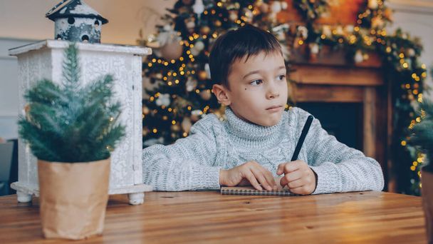 Portrait of thoughtful little boy writing letter to Santa Claus before Christmas in decorated room, child is sitting at table alone. New Year and holidays concept. - Фото, зображення