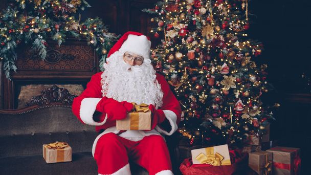 Santa is packing presents in bag getting ready for New Year celebrations near Christmas tree, decorations and fireplace are visible in background. - 写真・画像