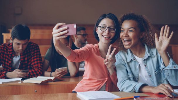 Cheerful girls students are taking selfie with smartphone sitting at tables at college, young women are posing making hand gestures and hugging. Friends and technology concept. - 写真・画像