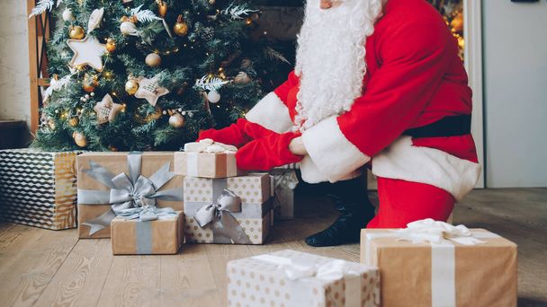 Santa Claus in traditional costume putting gift boxes under Christmas tree on holiday in house. Occasions and presents concept. - Photo, image
