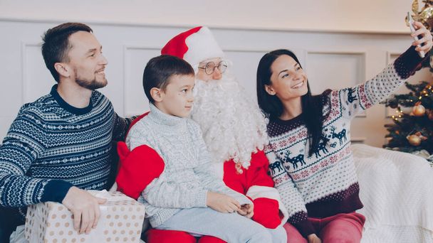 Happy young family mother, father and son taking selfie with Santa Claus using smartphone camera at home. Christmas, modern technology and people concept. - Fotografie, Obrázek