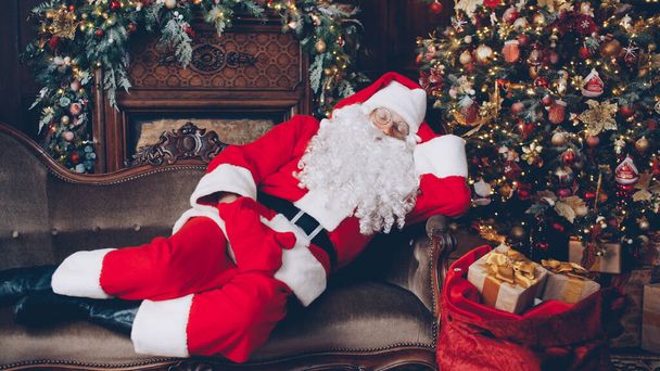 Santa Claus is sleeping in decorated room near Christmas tree with bag of gifts, man in costume is lying on sofa. Holidays and presents concept. - Fotografie, Obrázek