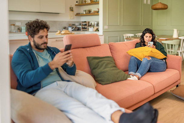 Young married couple using smart phone ignoring each other addicted to the internet and social network - man and woman phubbing in the living room - family people loneliness and technology lifestyle concept - Foto, imagen