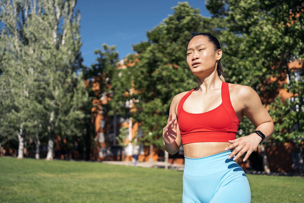 Silna Azjatka w stroju sportowym ćwicząca w parku w słoneczny dzień. Biegnie i patrzy przed siebie.. - Zdjęcie, obraz