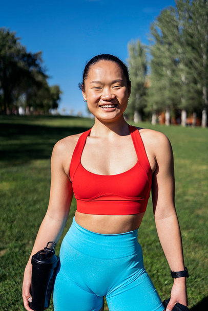 Strong asian woman with sports clothes working out in the park during sunny day. She is smiling and looking at camera. - Φωτογραφία, εικόνα