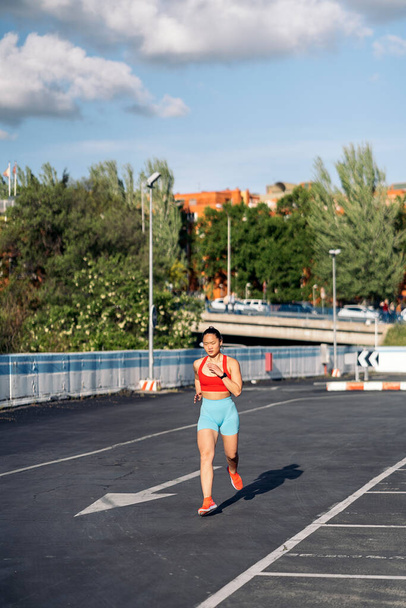 Strong asian woman with sports clothes working out in the street during sunny day. She is running and looking at front. - Fotografie, Obrázek