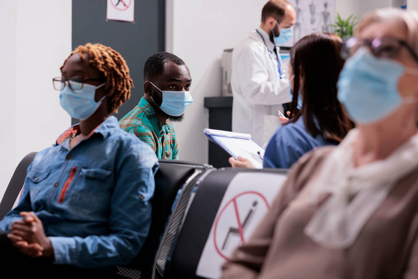 アフリカ系アメリカ人の患者は女性医師が通った。異なる民族、年齢の人々は、医師のオフィスの受付エリアでは、病院の待合室の一般的なショットでコビット19拡散を防止マスクを着用. - 写真・画像