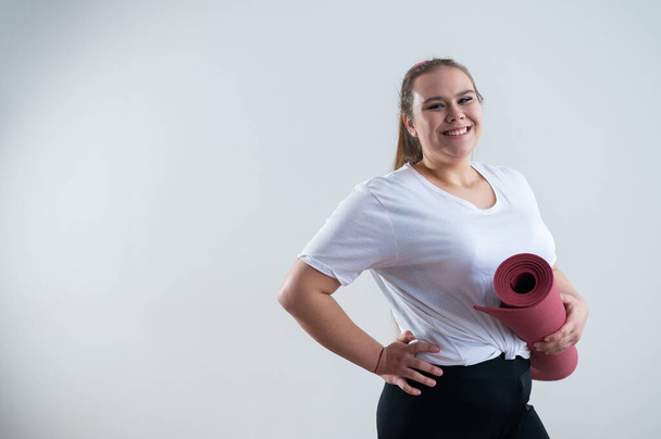 Young fat caucasian woman holding a sport mat. Charming plus size model in sportswear stands on a white background. - Foto, Imagem