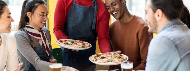 Baner poziomy lub nagłówek z kelnerem serwującym pyszne pizze margherita wielokulturowym przyjaciołom w przytulnej pizzerii - Multiethnic friends having fun together at the pizzeria - Zdjęcie, obraz