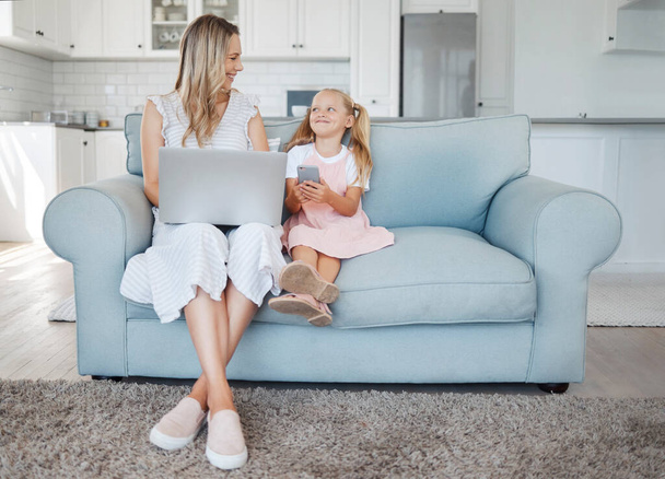 Mother, girl child and digital device happy, learn and smile for bonding, embrace and talking together in living room on couch. Laptop, smartphone and mama with daughter browse online and home school. - Фото, изображение