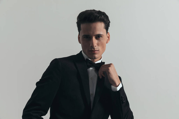 portrait of handsome young man in black tuxedo adjusting bowtie and posing in a confident way in front of grey background in studio - Foto, afbeelding