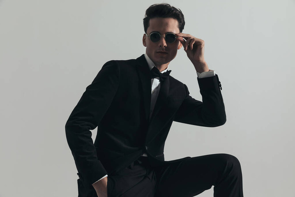 sexy uomo elegante con mano in tasca regolazione occhiali da sole e in posa con il ginocchio davanti a sfondo grigio in studio - Foto, immagini