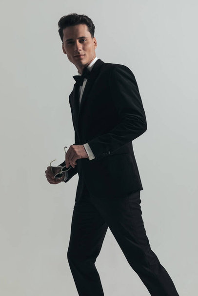side view of sexy man in black tuxedo holding sunglasses and smiling in front of grey background in studio - Фото, изображение