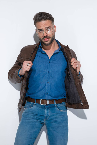 Sexy fashion model is arranging his brown leather jacket, posing with attitude against white studio background - Photo, Image