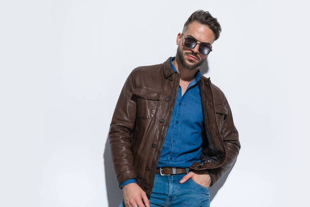 Sexy casual man putting one hand in pocket, wearing a brown leather jacket and sunglasses against white studio background - Фото, изображение