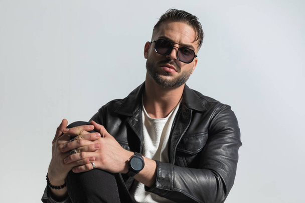 fashion man with wet hair wearing sunglasses and leather jacket and holding knee up with his hands while posing in front of white background in studio - Photo, Image
