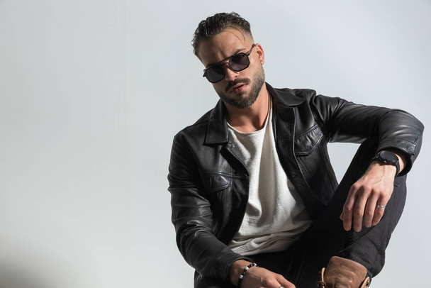 confident casual man wearing sunglasses and black leather jacket in a cool way, posing with his knee up and elbow above in front of grey background in studio, portrait - Photo, Image