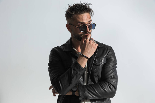 portrait of fashion man with messy wet hair wearing sunglasses and black leather jacket, crossing arms and looking away while sensually touching lips in studio - Photo, Image