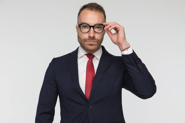 handsome businessman arranging his eyeglasses and wearing a nice suit and red tie on gray background - Photo, Image
