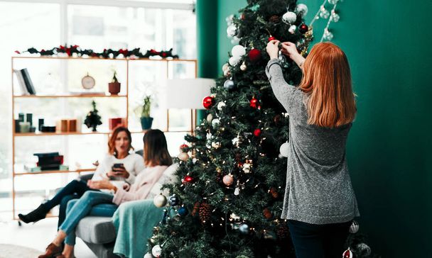 Ayudando con un poco de la decoración. Vista trasera de una mujer irreconocible decorando un árbol de Navidad en casa durante el día - Foto, Imagen