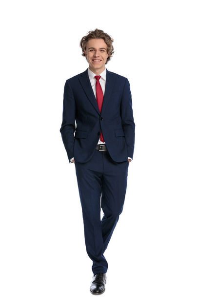 full body picture of happy young businessman in elegant suit holding hands in pockets, smiling and walking in front of white background in studio - Photo, Image