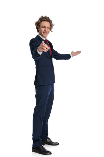 πλαϊνή άποψη εικόνα του σγουρά μαλλιά άνθρωπος στο κοστούμι με κόκκινη γραβάτα δείχνοντας τα δάχτυλα και καλώντας να πλευρά, ενώ στέκεται και ποζάρουν μπροστά από το λευκό φόντο, πλήρες σώμα - Φωτογραφία, εικόνα