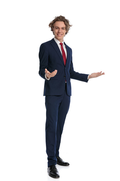 blonďatý mladý muž v elegantním obleku drží ruce a gestikuluje, usmívá se a prezentuje před bílým pozadím ve studiu, plné tělo - Fotografie, Obrázek