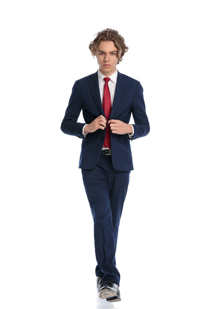 full length of sexy stylish businessman buttoning suit and walking in front of white background in studio, being confident and posing - Fotografie, Obrázek