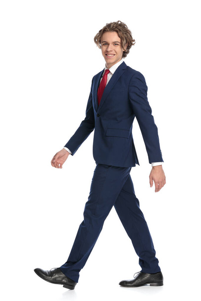 фотография сбоку элегантного бизнесмена в костюме с галстуком, улыбающегося и идущего перед белым фоном в студии, полное тело - Фото, изображение