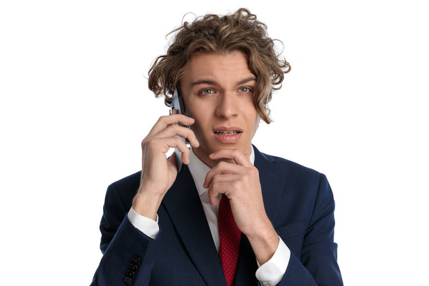 figyelmes férfi kék szemekkel és göndör hajjal, aki telefonon beszél és fehér háttér előtt gondolkodik a stúdióban, portré  - Fotó, kép