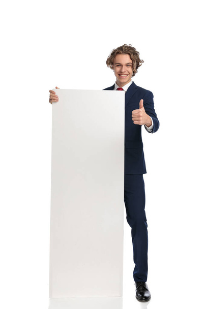 pohledný podnikatel v obleku prezentuje bílou prázdnou desku a usmívá se, zatímco dělá palce nahoru gesto před bílým pozadím ve studiu, plné tělo - Fotografie, Obrázek