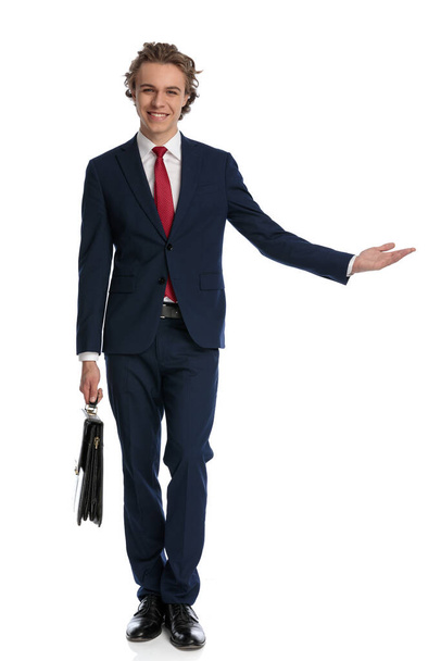 full body picture of happy businessman with suitcase smiling while inviting to side and showing in front of white background in studio - Zdjęcie, obraz
