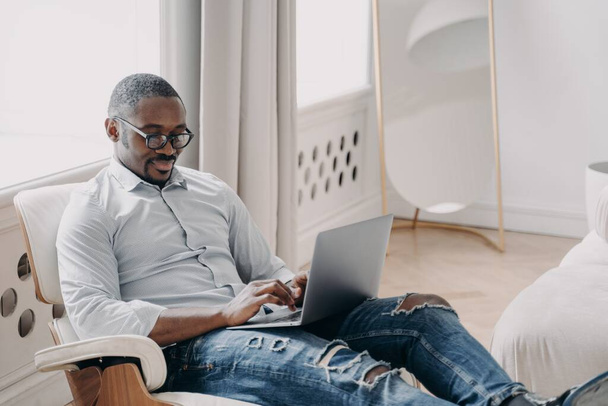 Freelancer afro-americano focado trabalhando online no projeto, sentado em poltrona com laptop. Homem de negócios negro moderno usando óculos digitando mensagem, respondendo e-mail. Conceito de trabalho remoto. - Foto, Imagem