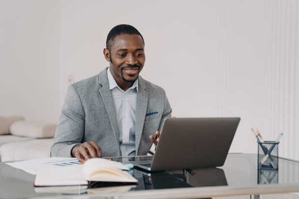 Hymyilevä afrikkalainen liikemies, joka on tyytyväinen bisnesprojektiinsa, työskentelee kannettavan tietokoneen parissa, lukee sähköpostia hyvillä uutisilla, istuu toimistossa. Musta mies työntekijä katselee webinaaria, oppiminen verkossa - Valokuva, kuva