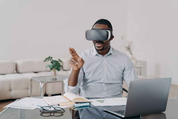 Αφροαμερικάνος σχεδιαστής αρχιτέκτονα που φοράει γυαλιά VR εργάζεται στο εσωτερικό του έργου σε εικονική πραγματικότητα, κάθεται στο γραφείο. Μοντέρνος μαύρος τύπος που χρησιμοποιεί υψηλής τεχνολογίας gadget για την εργασία στον κυβερνοχώρο. - Φωτογραφία, εικόνα