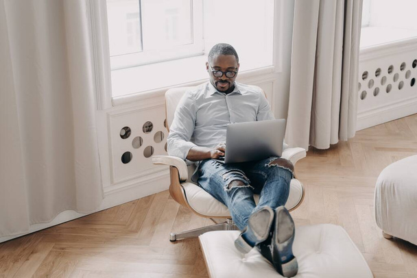 Skupiony afrykański biznesmen pracuje online na laptopie, siedzi w fotelu w domu. Nowoczesny czarny mężczyzna pracownik menedżer w okularach odpowiada e-mail, rozwój projektu biznesowego. Koncepcja pracy na odległość. - Zdjęcie, obraz