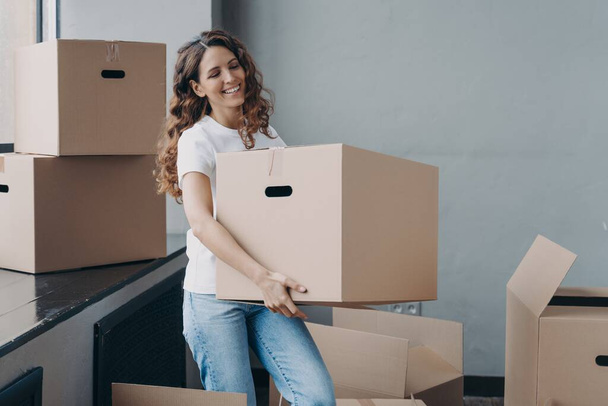 Moving day. Happy female tenant homeowner carrying cardboard box with household things. Smiling girl enjoying preparations to relocation to new house, packing belongings. Removal and delivery service. - Foto, immagini