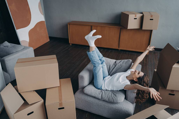 Happy young woman celebrating relocation to first new home, having fun packing, raising hands sitting in armchair on day of moving, excited girl do yes gesture rejoicing surrounded by cardboard boxes. - Photo, Image