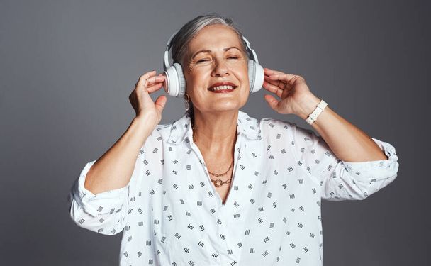 Můžu poslouchat rozkošnou hudbu na cestách. Studio záběr starší ženy na sluchátka při poslechu hudby na šedém pozadí - Fotografie, Obrázek