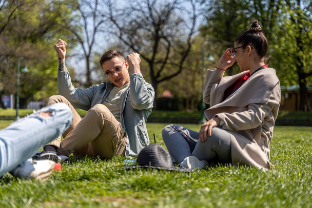 Group of cheerful students teenagers in casual outfits sitting on a green grass in park, enjoying - Photo, Image