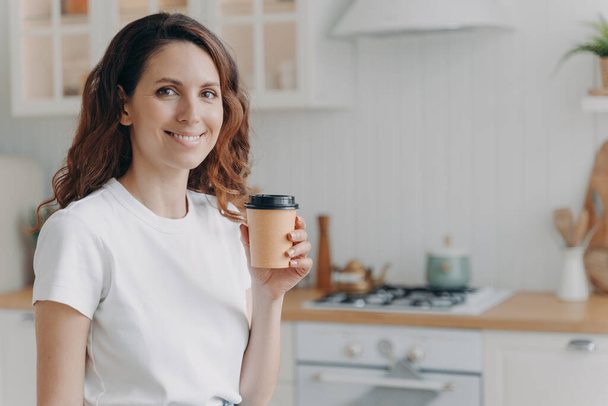 Portré barátságos boldog spanyol nő kezében eldobható papír csésze kávét a modern konyhában. Mosolygó nyugodt női háziasszony tart szünetet házimunka közben, élvezi meleg ital otthon.  - Fotó, kép