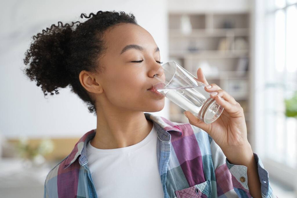 Zijaanzicht van jonge gemengde ras meisje drinken schoon mineraal stilstaand water uit glas thuis. Mooie dorstige tienerdame genieten van dagelijkse gezonde levensstijl gewoonte. Natuurlijke schoonheid, lichaamsverzorging, wellness. - Foto, afbeelding