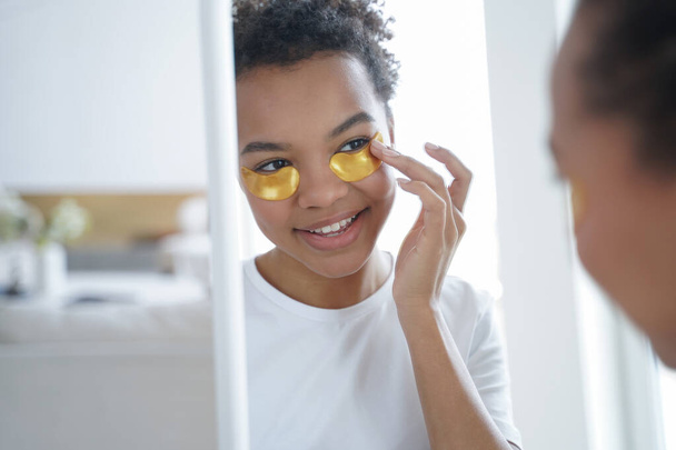 Pretty mixed race girl caring for skin applying golden under eye hydrogel patches, looking in the mirror, smiling young lady uses collagen pads for skincare. Daily beauty habit, cosmetics advertising. - Photo, Image