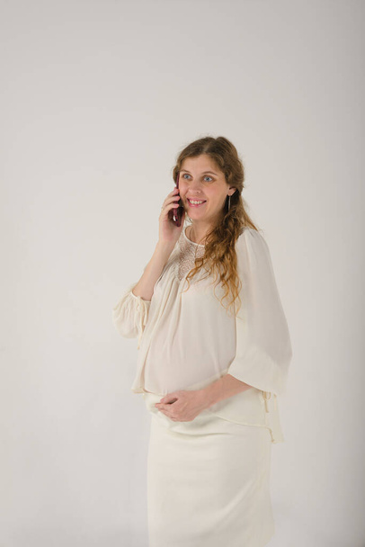 portrait of a pregnant woman in a business white blouse and skirt, talking on the phone, on a light plain background - Foto, imagen