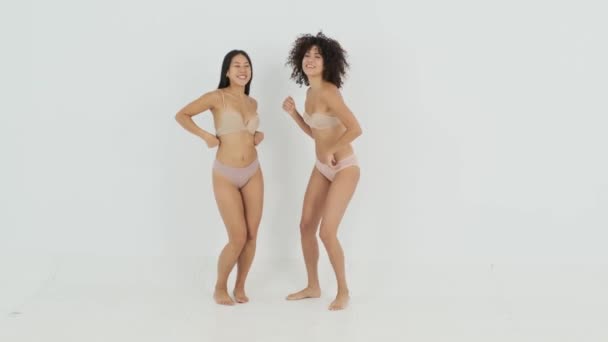 Pełne treści młodych wielonarodowych modeli kobiet w wygodnej bieliźnie z długimi ciemnymi włosami uśmiechających się szczęśliwie podczas tańca na białym tle w studio - Materiał filmowy, wideo