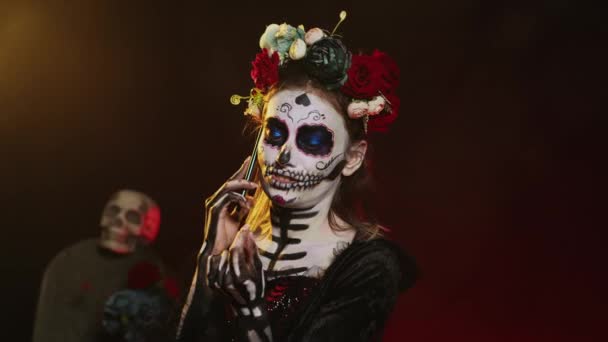 Lady of dead talking on phone call with smartphone, wearing flowers crown and cavalera catrina halloween costume. Woman dressed as santa muerte to celebrate mexican culture holiday. Handheld shot. - Footage, Video
