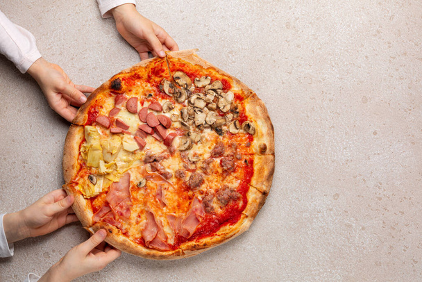Girl's hands with pizza Four seasons. Mozzarella cheese, tomatoes,  mushrooms, sausage, prosciutto, Italian made pizza Margherita on beige concrete background. Italy. Top view. Copy space. - 写真・画像